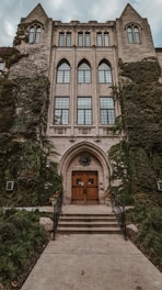 A historic, ivy-covered building features tall, arched windows and a grand entrance with wooden doors. The facade is adorned with intricate stonework and is framed by metal railings leading to the entrance.