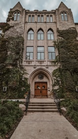 A historic, ivy-covered building features tall, arched windows and a grand entrance with wooden doors. The facade is adorned with intricate stonework and is framed by metal railings leading to the entrance.