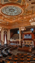 An ornate legislative chamber with elaborate design elements featuring a grand stained glass dome ceiling, gold and bronze accents, and multiple chandeliers. The room is filled with rows of curved wooden desks and leather chairs, surrounded by Corinthian columns and large draped windows. A speaker's podium and electronic displays are positioned at the front.