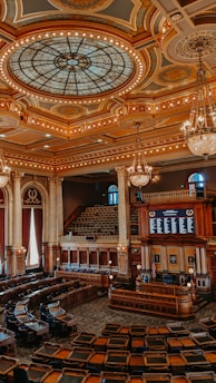 An ornate legislative chamber with elaborate design elements featuring a grand stained glass dome ceiling, gold and bronze accents, and multiple chandeliers. The room is filled with rows of curved wooden desks and leather chairs, surrounded by Corinthian columns and large draped windows. A speaker's podium and electronic displays are positioned at the front.