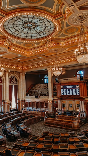 An ornate legislative chamber with elaborate design elements featuring a grand stained glass dome ceiling, gold and bronze accents, and multiple chandeliers. The room is filled with rows of curved wooden desks and leather chairs, surrounded by Corinthian columns and large draped windows. A speaker's podium and electronic displays are positioned at the front.