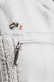 A close-up of a textured white jacket featuring a zippered pocket. On the jacket's surface, there's a small enamel pin with the design of two crossed flags in red, green, black, and white colors.