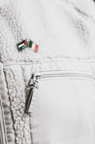 A close-up of a textured white jacket featuring a zippered pocket. On the jacket's surface, there's a small enamel pin with the design of two crossed flags in red, green, black, and white colors.