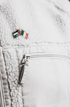 A close-up of a textured white jacket featuring a zippered pocket. On the jacket's surface, there's a small enamel pin with the design of two crossed flags in red, green, black, and white colors.