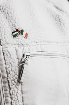 A close-up of a textured white jacket featuring a zippered pocket. On the jacket's surface, there's a small enamel pin with the design of two crossed flags in red, green, black, and white colors.
