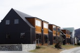 A row of modern townhouses featuring a combination of black and wooden exteriors. The buildings have large windows and sit on a foundation of stone walls. There is a tree and some bushes in a grassy area in front of the houses, and a road runs alongside the buildings.