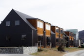 A row of modern townhouses featuring a combination of black and wooden exteriors. The buildings have large windows and sit on a foundation of stone walls. There is a tree and some bushes in a grassy area in front of the houses, and a road runs alongside the buildings.