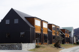 A row of modern townhouses featuring a combination of black and wooden exteriors. The buildings have large windows and sit on a foundation of stone walls. There is a tree and some bushes in a grassy area in front of the houses, and a road runs alongside the buildings.