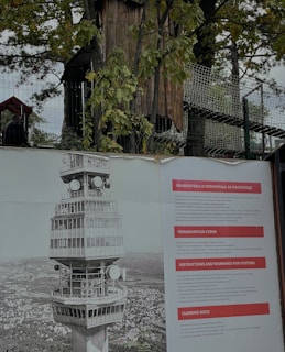 A large poster is displayed outdoors featuring an illustration of a tall, distinctive tower with multiple levels and satellite dishes. Beside the illustration, there are informational panels in multiple languages, including English, with titles like 'Instructions and Warnings for Visitors' and 'Climbing Rock'. Behind the poster, a fenced area with trees and some wooden structures is visible.