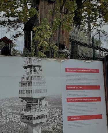 A large poster is displayed outdoors featuring an illustration of a tall, distinctive tower with multiple levels and satellite dishes. Beside the illustration, there are informational panels in multiple languages, including English, with titles like 'Instructions and Warnings for Visitors' and 'Climbing Rock'. Behind the poster, a fenced area with trees and some wooden structures is visible.