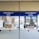 Two service windows with blue signs above each window. The left sign reads 'Facturacion Caja' and the right one reads 'Faturamento Caixa.' These windows appear to be part of a billing or payment service area. A few items, such as boxes and papers, are placed behind the windows. A small digital display shows a number.