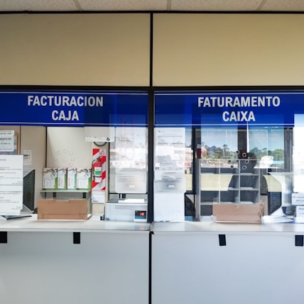 Two service windows with blue signs above each window. The left sign reads 'Facturacion Caja' and the right one reads 'Faturamento Caixa.' These windows appear to be part of a billing or payment service area. A few items, such as boxes and papers, are placed behind the windows. A small digital display shows a number.
