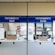 Two service windows with blue signs above each window. The left sign reads 'Facturacion Caja' and the right one reads 'Faturamento Caixa.' These windows appear to be part of a billing or payment service area. A few items, such as boxes and papers, are placed behind the windows. A small digital display shows a number.