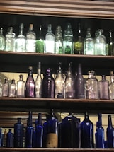 A collection of antique bottles arranged on wooden shelves. The bottles vary in size, color, and shape, with some made of clear or lightly tinted glass, while others are deeper shades of green, purple, and blue. The lighting emphasizes the transparency and colors of the glass.