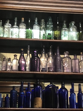 A collection of antique bottles arranged on wooden shelves. The bottles vary in size, color, and shape, with some made of clear or lightly tinted glass, while others are deeper shades of green, purple, and blue. The lighting emphasizes the transparency and colors of the glass.