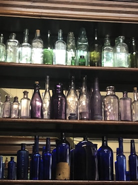 A collection of antique bottles arranged on wooden shelves. The bottles vary in size, color, and shape, with some made of clear or lightly tinted glass, while others are deeper shades of green, purple, and blue. The lighting emphasizes the transparency and colors of the glass.