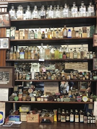 Shelves stocked with vintage apothecary bottles and containers, featuring a variety of old medicinal liquids and powders. Some bottles have handwritten or printed labels displaying names of chemicals. The display includes various sizes and shapes of bottles, with a mix of glass and metal containers. Visible behind the shelves is a mirror reflecting parts of the shop and additional items.