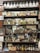 Shelves stocked with vintage apothecary bottles and containers, featuring a variety of old medicinal liquids and powders. Some bottles have handwritten or printed labels displaying names of chemicals. The display includes various sizes and shapes of bottles, with a mix of glass and metal containers. Visible behind the shelves is a mirror reflecting parts of the shop and additional items.