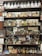 Shelves stocked with vintage apothecary bottles and containers, featuring a variety of old medicinal liquids and powders. Some bottles have handwritten or printed labels displaying names of chemicals. The display includes various sizes and shapes of bottles, with a mix of glass and metal containers. Visible behind the shelves is a mirror reflecting parts of the shop and additional items.