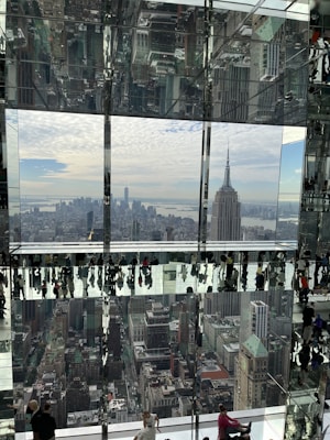 A modern urban scene from a high vantage point features a reflective glass interior with numerous people walking and interacting. The cityscape in the background includes prominent skyscrapers under a cloudy sky, with reflections amplifying the architectural complexity.