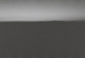 A monochrome landscape featuring a dark, shadowy foreground with a distant horizon line separating the land from the gradient sky. The overall texture appears grainy, giving it a vintage or film-like quality.