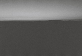 A monochrome landscape featuring a dark, shadowy foreground with a distant horizon line separating the land from the gradient sky. The overall texture appears grainy, giving it a vintage or film-like quality.