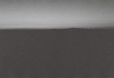 A monochrome landscape featuring a dark, shadowy foreground with a distant horizon line separating the land from the gradient sky. The overall texture appears grainy, giving it a vintage or film-like quality.