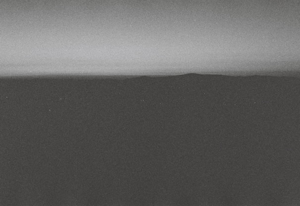 A monochrome landscape featuring a dark, shadowy foreground with a distant horizon line separating the land from the gradient sky. The overall texture appears grainy, giving it a vintage or film-like quality.