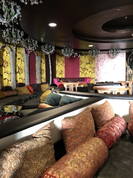 A richly decorated interior space featuring vibrant yellow floral-patterned walls, plush seating with a mix of patterned cushions in red, pink, and gold, and ornate chandeliers hanging from the ceiling. The room includes tables and has a luxurious, eclectic style with a mix of textures and elegant decor elements.