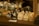 A collection of small, handcrafted wooden cat figurines with different expressions and colors displayed on a wooden shelf. The background includes a rack with various decorative items and a wicker basket. A handwritten label in the foreground indicates these are carved cat figures.