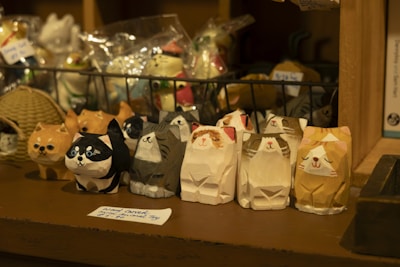 A collection of small, handcrafted wooden cat figurines with different expressions and colors displayed on a wooden shelf. The background includes a rack with various decorative items and a wicker basket. A handwritten label in the foreground indicates these are carved cat figures.
