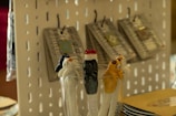 Ceramic cat-shaped utensils rest in a glass, featuring varying designs and colors. In the background, products hang on hooks against a perforated display board.
