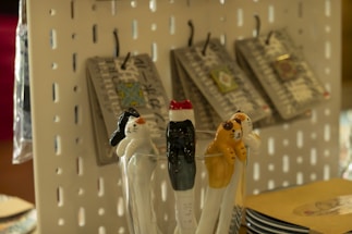 Ceramic cat-shaped utensils rest in a glass, featuring varying designs and colors. In the background, products hang on hooks against a perforated display board.