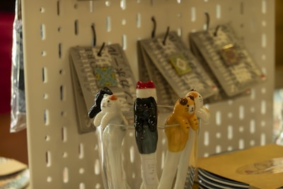 Ceramic cat-shaped utensils rest in a glass, featuring varying designs and colors. In the background, products hang on hooks against a perforated display board.