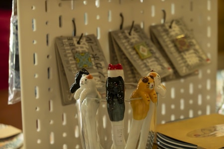 Ceramic cat-shaped utensils rest in a glass, featuring varying designs and colors. In the background, products hang on hooks against a perforated display board.