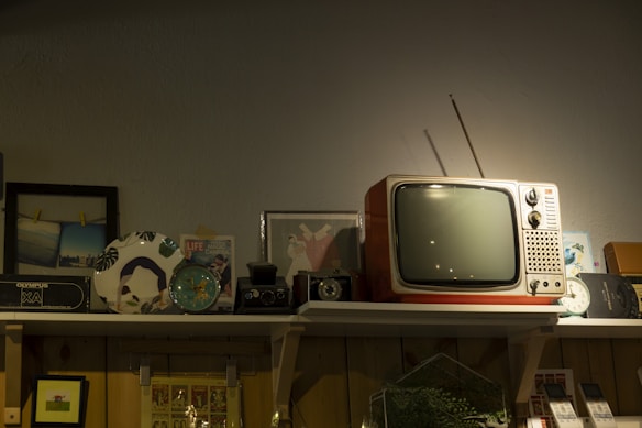 A vintage scene featuring an old-fashioned television with antennas sits prominently on a shelf, surrounded by various retro items. These include an analog clock, a classic camera, framed pictures, and decorative plates. The warm lighting casts a nostalgic ambiance over the assemblage.