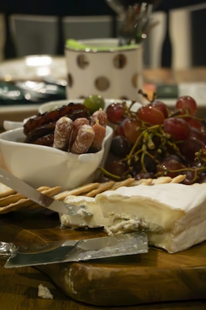 A platter featuring a block of soft, creamy cheese with a piece freshly cut and knives placed on top. Next to the cheese are cracker sticks, a bunch of grapes, and a small bowl containing cured salami sticks. The background is softly blurred, with a container that has circular patterns.