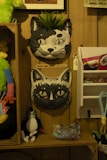 The image features a display of various cat-themed items on wooden shelves and background. There are two decorative cat vases mounted on the wall, one with leaves coming out of the top, resembling a plant. Below the vases, stuffed cat toys and other cat-themed merchandise are arranged neatly.