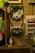 The image features a display of various cat-themed items on wooden shelves and background. There are two decorative cat vases mounted on the wall, one with leaves coming out of the top, resembling a plant. Below the vases, stuffed cat toys and other cat-themed merchandise are arranged neatly.