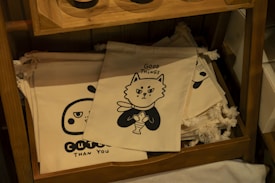 The image contains a wooden shelf displaying several cloth bags with printed designs. One of the prominent bags features a cartoon cat holding a fish with the text 'Good Things', and another shows a cute face with the text 'Cuter Than You'. The bags are stacked neatly.
