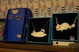 Several jewelry items are displayed on a wooden surface. There are two blue boxes with necklaces featuring cat-shaped pendants. One of the pendants depicts a cat wearing a small hat, while the other depicts a cat with a red bow. Next to these is another blue box featuring a pair of 24K gold plated earrings shaped like cat faces, priced at $89. The background consists of a patterned wall with stylized designs.