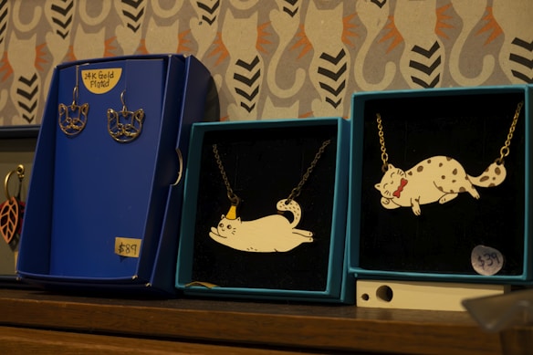 Several jewelry items are displayed on a wooden surface. There are two blue boxes with necklaces featuring cat-shaped pendants. One of the pendants depicts a cat wearing a small hat, while the other depicts a cat with a red bow. Next to these is another blue box featuring a pair of 24K gold plated earrings shaped like cat faces, priced at $89. The background consists of a patterned wall with stylized designs.