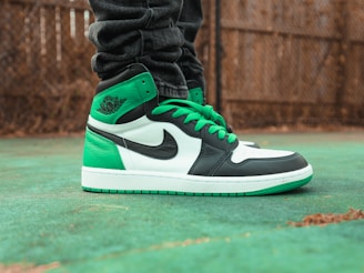 A close-up of a stylish green, white, and black sneaker with green laces on a green outdoor surface. The sneaker is worn by a person dressed in dark, rolled-up jeans. A blurred background featuring a brown wooden fence adds depth to the scene.