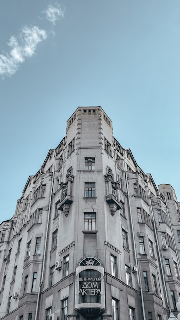 A tall, ornate building with a classical architectural style stands prominently against a clear blue sky. The facade features a combination of vertical lines and decorative elements, with multiple windows arranged symmetrically. At the base of the building, there is a sign with text in a foreign language.