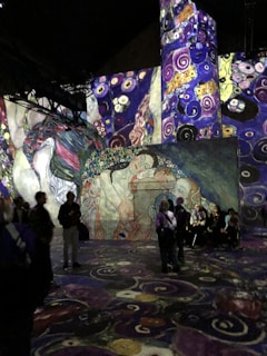 A large, immersive art installation featuring vibrant, swirling patterns inspired by Gustav Klimt's style. The image shows a room with walls and floors adorned with intricate designs, including representations of human figures and decorative floral motifs. Several people are walking or standing, observing the projections.