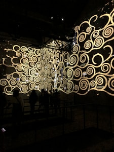 A dark room features intricate golden spirals and swirling patterns projected onto the walls. These ornate designs resemble an art installation or digital projection. Silhouettes of people can be seen observing the display, adding a sense of scale and engagement.