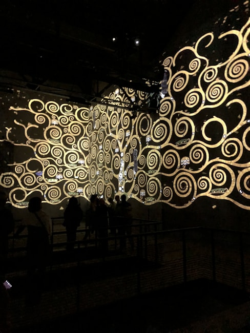 A dark room features intricate golden spirals and swirling patterns projected onto the walls. These ornate designs resemble an art installation or digital projection. Silhouettes of people can be seen observing the display, adding a sense of scale and engagement.