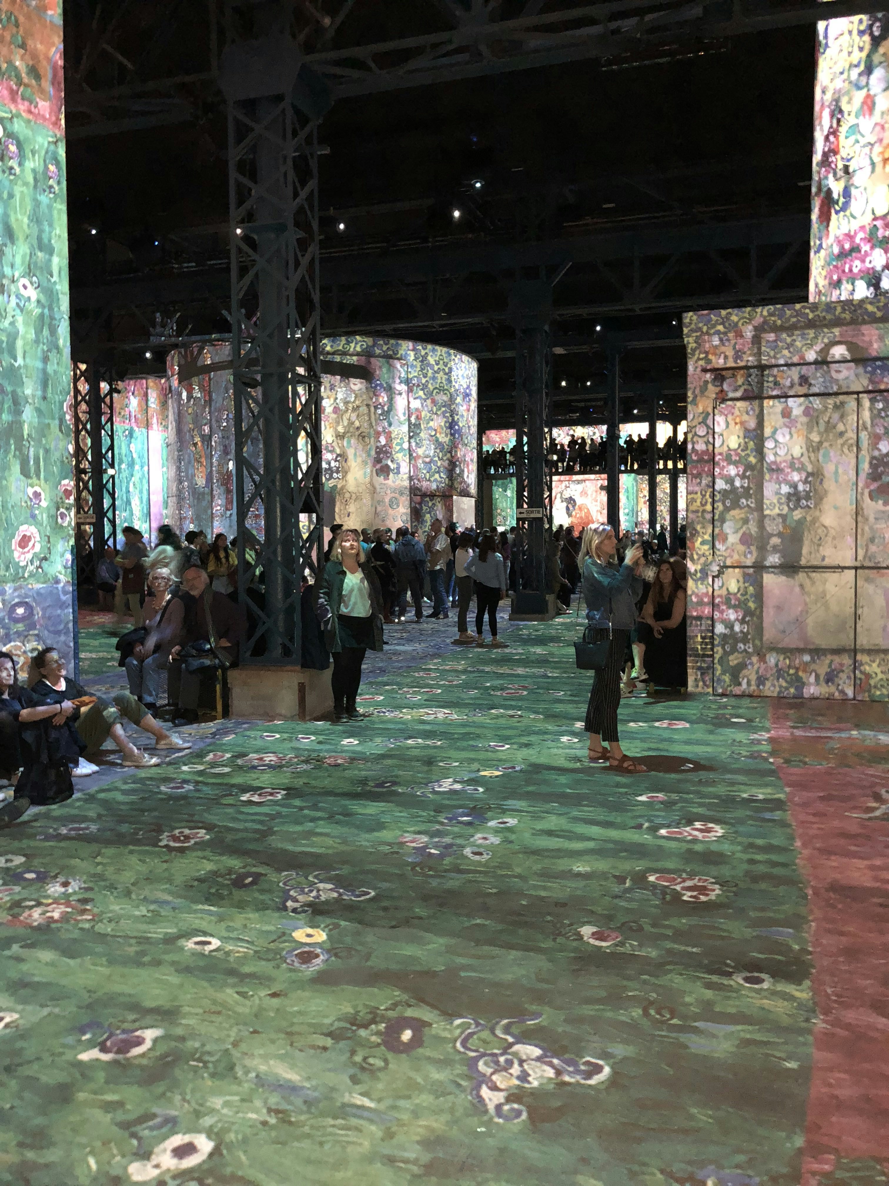 A large indoor exhibition space with projections of colorful floral and abstract patterns on walls and floors. The room is filled with people, some sitting and others walking, engaging with the immersive art experience. Tall metal columns are present, supporting the structure.