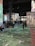 A large indoor exhibition space with projections of colorful floral and abstract patterns on walls and floors. The room is filled with people, some sitting and others walking, engaging with the immersive art experience. Tall metal columns are present, supporting the structure.