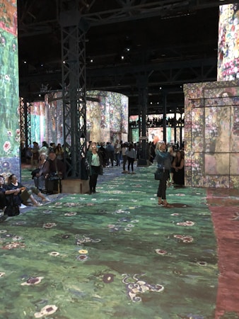 A large indoor exhibition space with projections of colorful floral and abstract patterns on walls and floors. The room is filled with people, some sitting and others walking, engaging with the immersive art experience. Tall metal columns are present, supporting the structure.